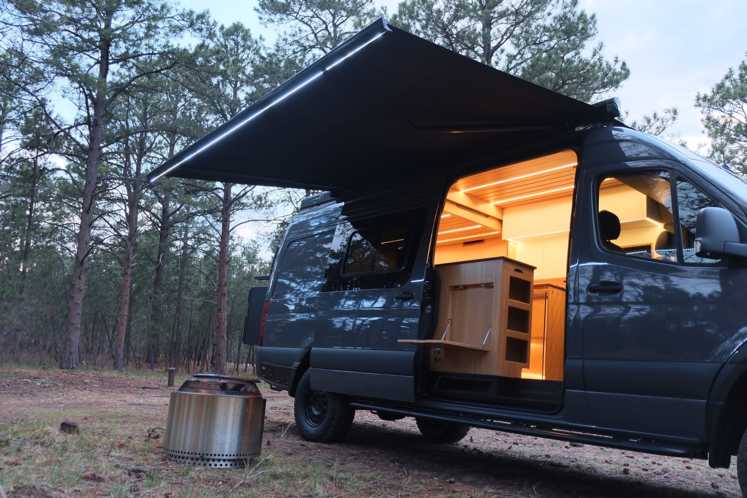 The Den Sprinter camper in a pine forest with its awning extended and warmly lit side door open.