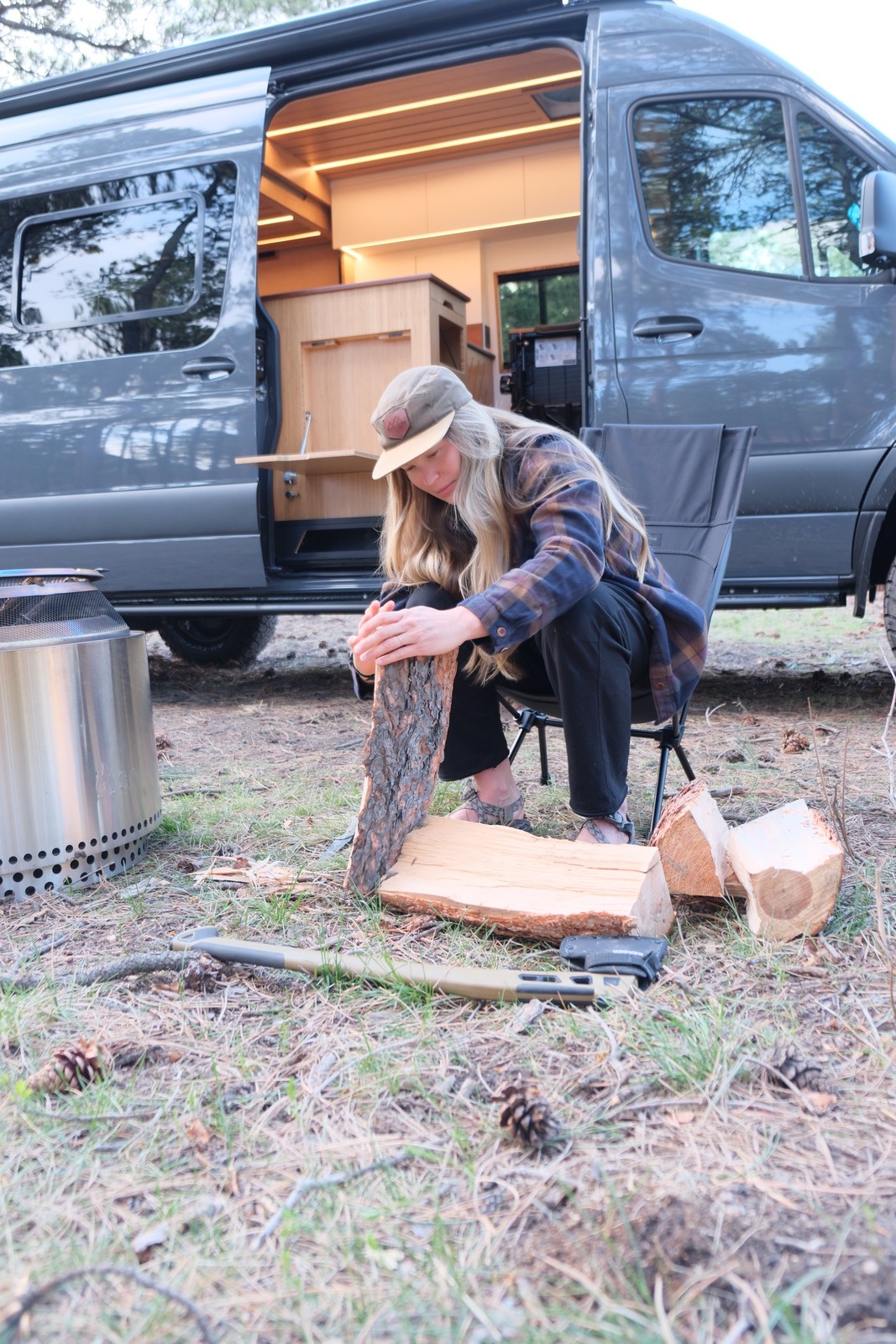 A person preparing firewood beside the Den open side door at a forest campsite.