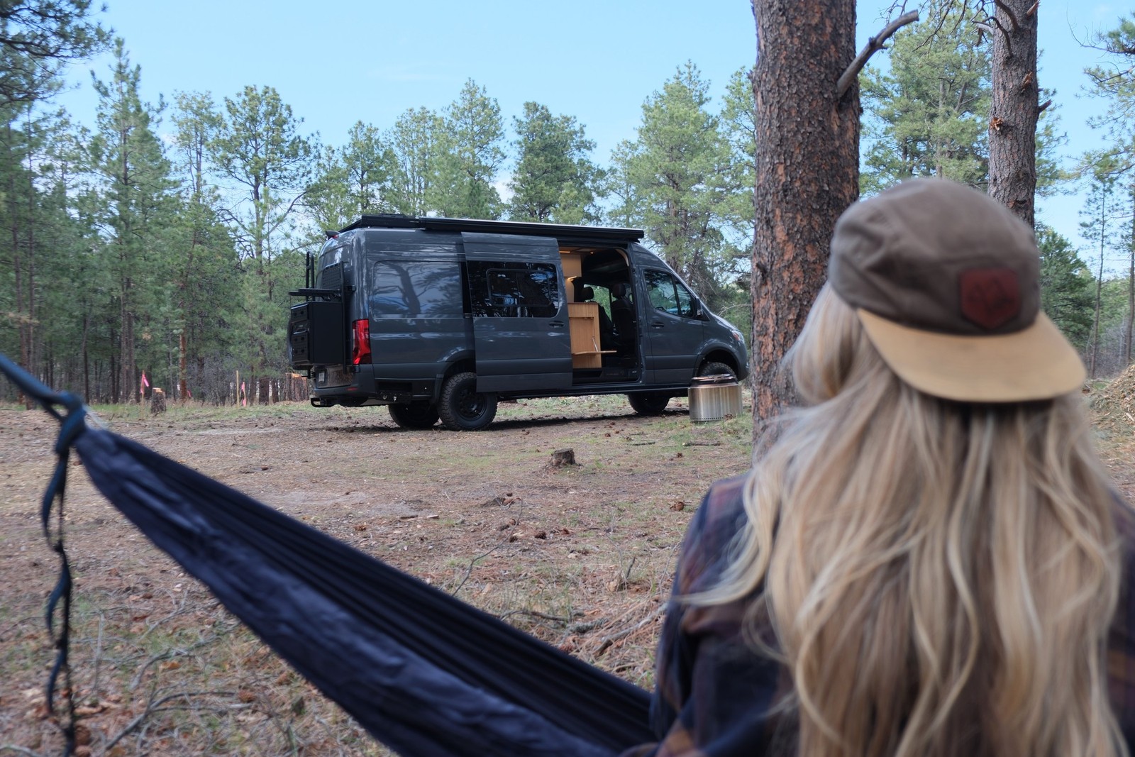 The Den parked in the woods with a person relaxing in a hammock in the foreground.